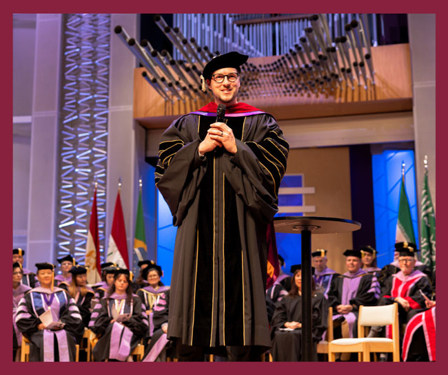 Dedication speaker Pastor Filip Milpsavljevic standing at a podium in academic regalia, addressing new students and faculty during a university dedication ceremony
