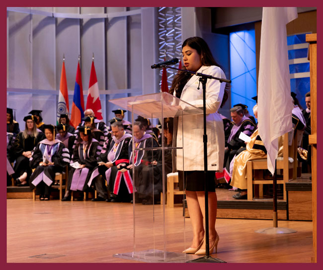 Student speaker in a white coat stands at a podium during a School of Dentistry dedication ceremony, with faculty members seated on stage in the background