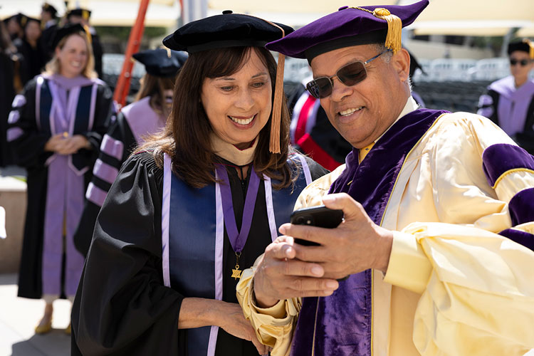 Ronald Forde and Esther Forde wearing academic regalia standing outdoors and looking at a smartphone before a hooding ceremony