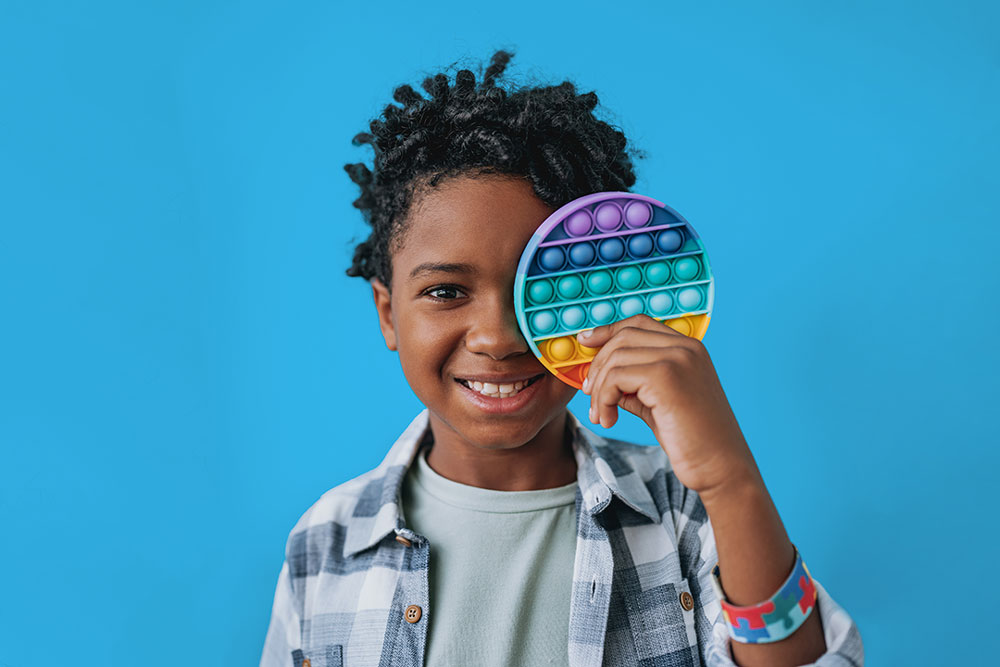 Child holding a rainbow pop‑it sensory toy over one eye against a blue background