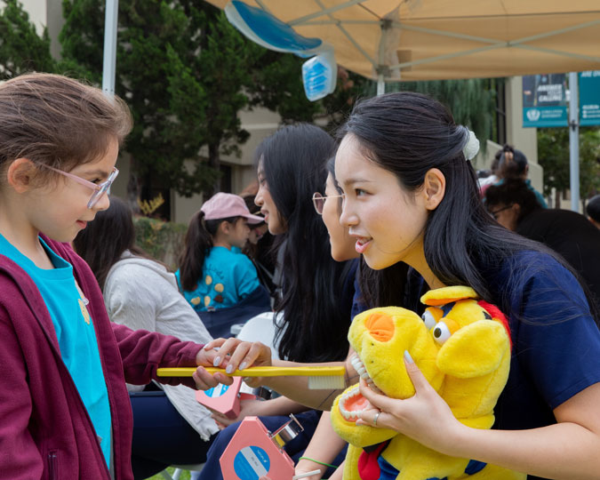 Child receiving hands-on oral hygiene education with a large toothbrush and dental model at a community children’s health fair, with volunteers and families in the background.