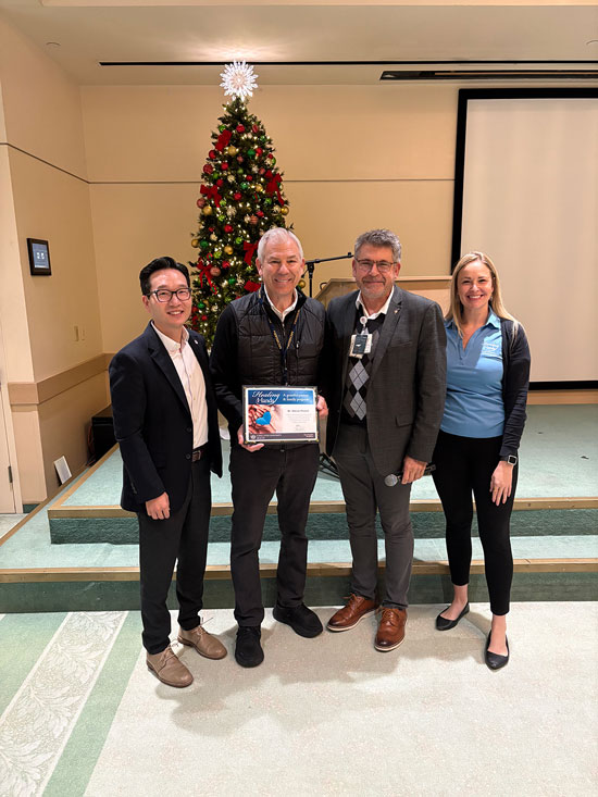 John Won, Steven Powell, Tim Sherwin, and Lauren Oliphant standing indoors in front of a decorated Christmas tree, with Steven Powell holding an Healing Hand certificate