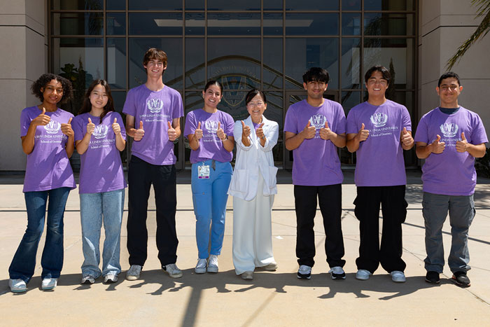 Group of people standing outdoors in front of a building, wearing matching purple shirts and giving thumbs up