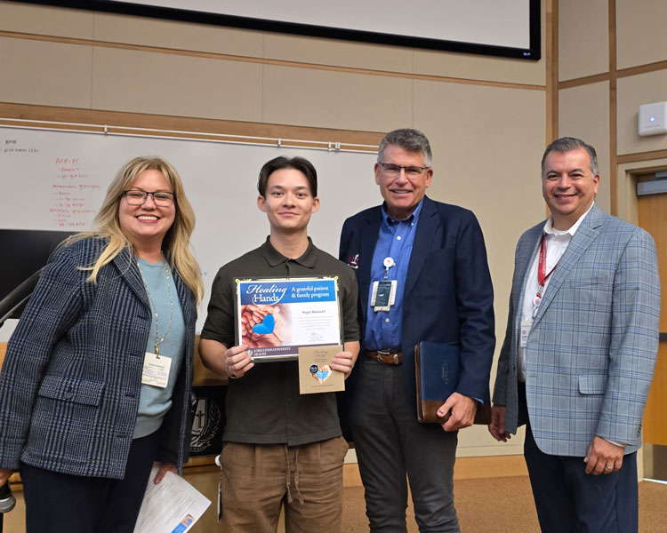 Four adults standing in a classroom setting, with Nigel holding a “Healing Hands” certificate and a small gift bag during an award presentation. A whiteboard with writing is visible in the background, and all individuals are facing the camera.