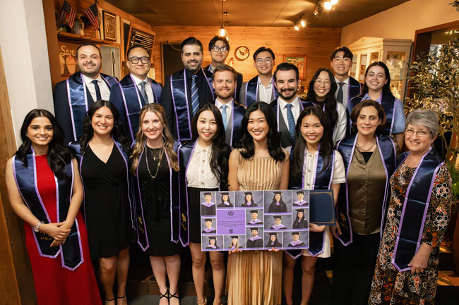 Large group of university students, faculty, and staff wearing purple graduation stoles posing indoors at an honor society banquet, with framed photos of members displayed in front.
