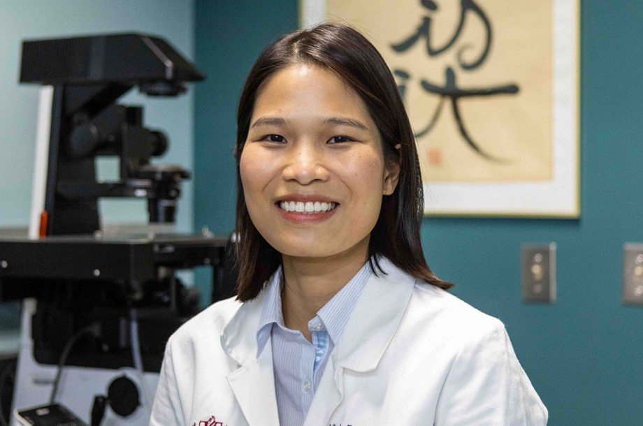 Dr. Minh Phuong Dang smiles in a Loma Linda University lab coat, seated beside a microscope in a research lab.
