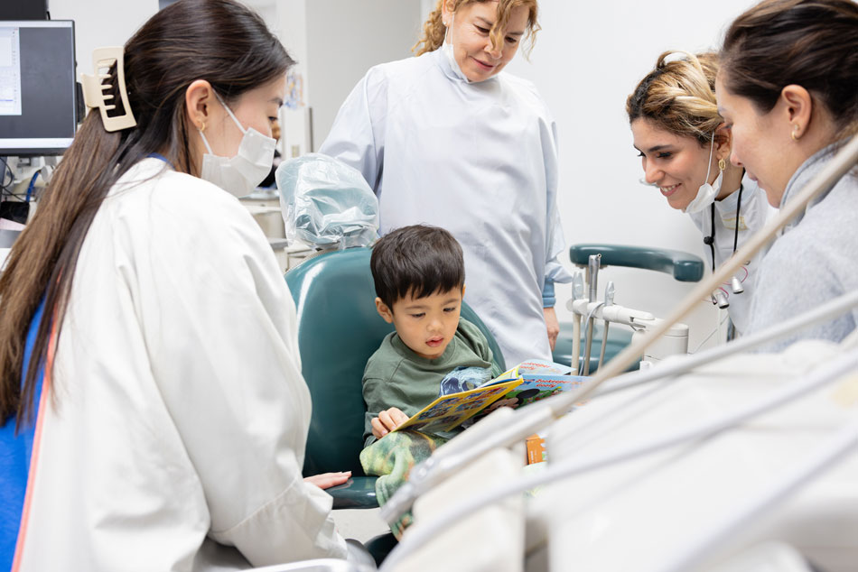 Child sitting in dental chair reading a book while a team of dental professionals provide pediatric dental care in a clinic.