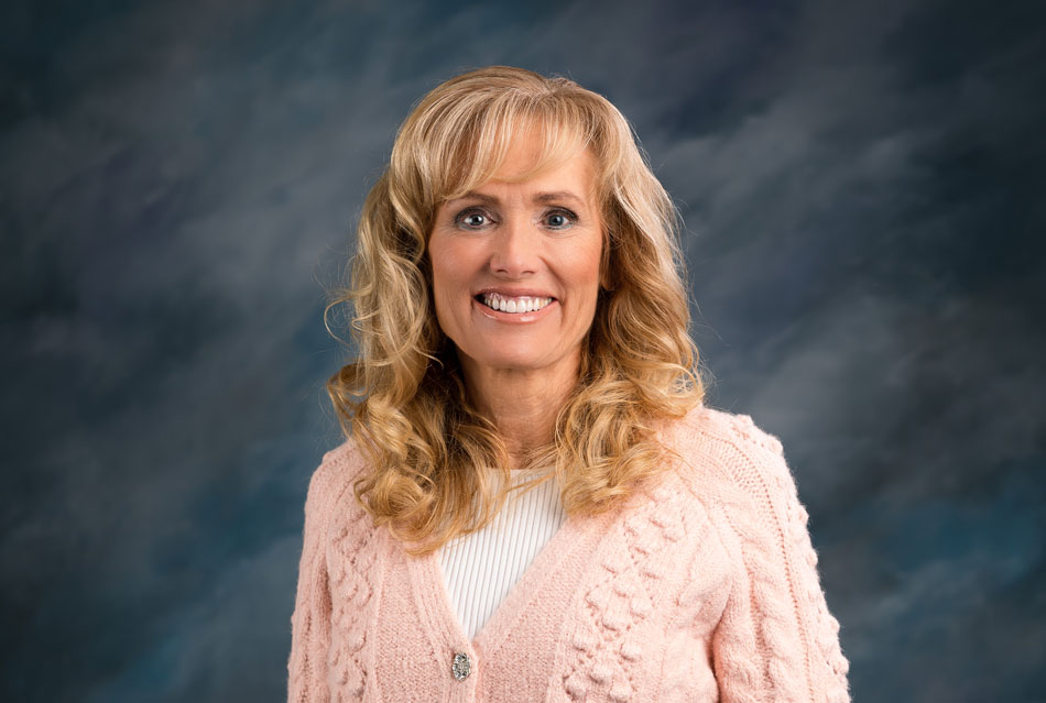 Professional headshot of Shelley Hayton smiling wearing a pink textured cardigan and white top, photographed in a studio with a blue gradient background
