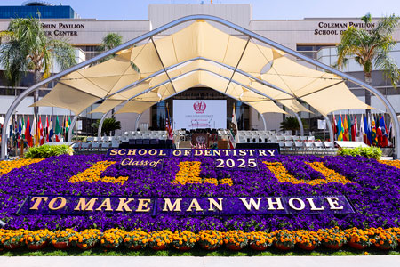 Outdoor commencement stage with floral display reading "School of Dentistry Class of 2025 To Make Man Whole" in front of a pavilion
