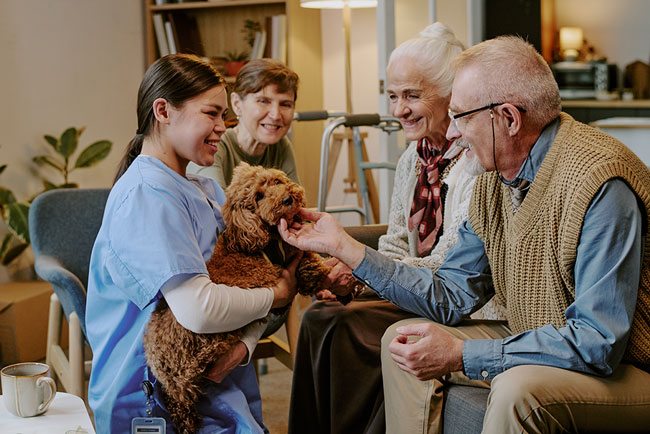 A dental hygiene professional introduces a therapy dog to two elderly patients to help reduce dental anxiety during a visit.