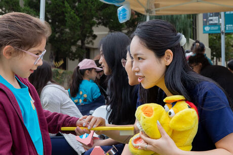  Child receiving hands-on oral hygiene education with a large toothbrush and dental model at a community children’s health fair, with volunteers and families in the background.