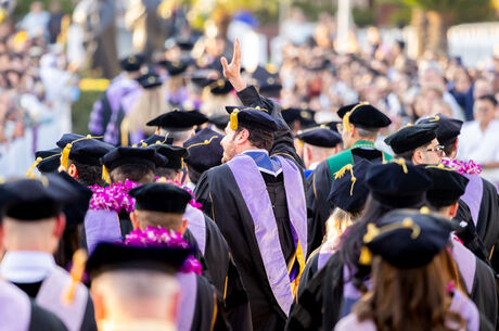 Group of LLUSD graduates in caps and gowns at an outdoor commencement ceremony, with one graduate raising a hand.