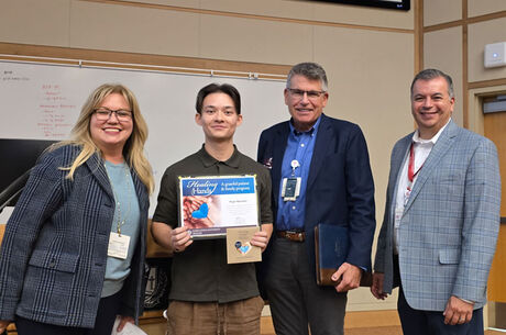 Four adults standing in a classroom setting, with Nigel holding a “Healing Hands” certificate and a small gift bag during an award presentation. A whiteboard with writing is visible in the background, and all individuals are facing the camera.