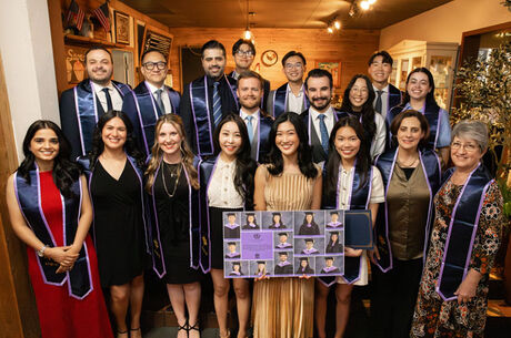 Large group of university students, faculty, and staff wearing purple graduation stoles posing indoors at an honor society banquet, with framed photos of members displayed in front.