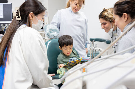 Child sitting in dental chair reading a book while a team of dental professionals provide pediatric dental care in a clinic.