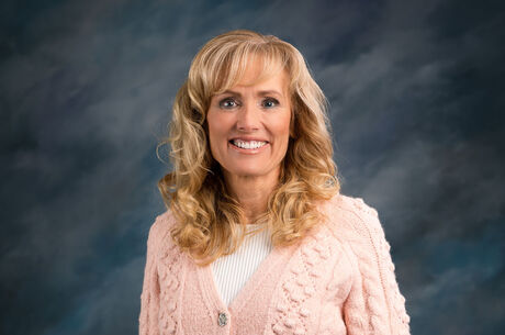 Professional headshot of Shelley Hayton smiling wearing a pink textured cardigan and white top, photographed in a studio with a blue gradient background