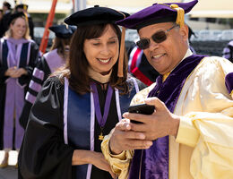 Ronald Forde and Esther Forde wearing academic regalia standing outdoors and looking at a smartphone before a hooding ceremony