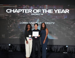 Three people standing on a stage holding a certificate in front of a screen reading "Chapter of the Year, District 13, Loma Linda University"