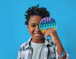 Child holding a rainbow pop‑it sensory toy over one eye against a blue background