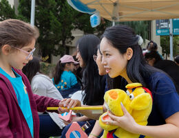  Child receiving hands-on oral hygiene education with a large toothbrush and dental model at a community children’s health fair, with volunteers and families in the background.