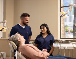 Dental care professionals speaking with a patient seated in a dental chair inside a treatment room.