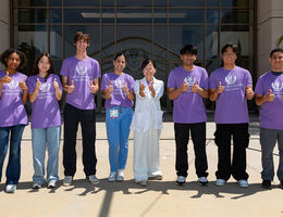 Group of people standing outdoors in front of a building, wearing matching purple shirts and giving thumbs up