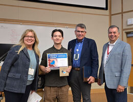 Four adults standing in a classroom setting, with Nigel holding a “Healing Hands” certificate and a small gift bag during an award presentation. A whiteboard with writing is visible in the background, and all individuals are facing the camera.