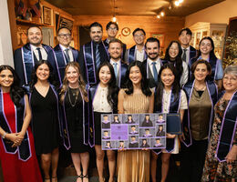 Large group of university students, faculty, and staff wearing purple graduation stoles posing indoors at an honor society banquet, with framed photos of members displayed in front.