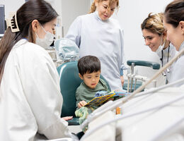 Child sitting in dental chair reading a book while a team of dental professionals provide pediatric dental care in a clinic.
