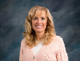 Professional headshot of Shelley Hayton smiling wearing a pink textured cardigan and white top, photographed in a studio with a blue gradient background